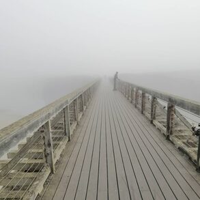 On a bridge in the fog a man in sillhouette looks over the railing